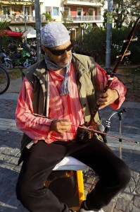 A man playing a traditional string instrument while sitting outdoors, wearing a patterned headscarf and sunglasses.